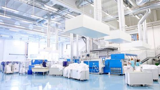Image of building interior with commercial laundry system, uniformed workers utilizing the machines, and clean linens stacked in rolling carts.